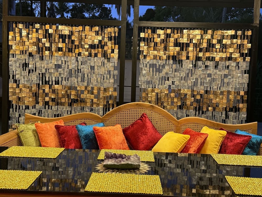 A vibrant outdoor lounge area features colorful cushions on wicker chairs, a reflective table adorned with golden accents, and shimmering mosaic walls, creating an inviting and luxurious atmosphere perfect for a tropical getaway.