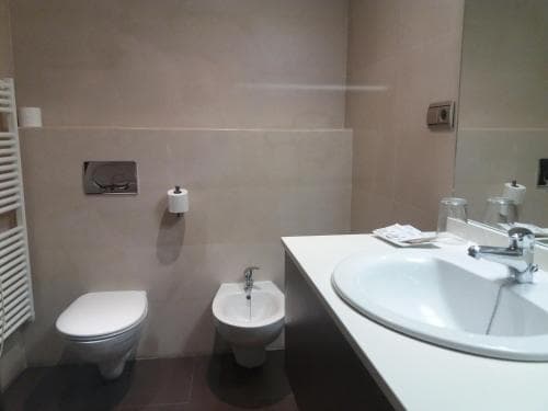 A modern bathroom with beige tiled walls, a sleek white sink, toilet, bidet, and towel radiator exudes a clean and minimalist vibe, ideal for travelers seeking comfort in accommodations.