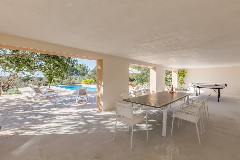 A serene outdoor patio features a dining table, lounge chairs, and a ping pong table under a shaded canopy, overlooking a tranquil pool surrounded by lush greenery, ideal for a relaxing vacation retreat.