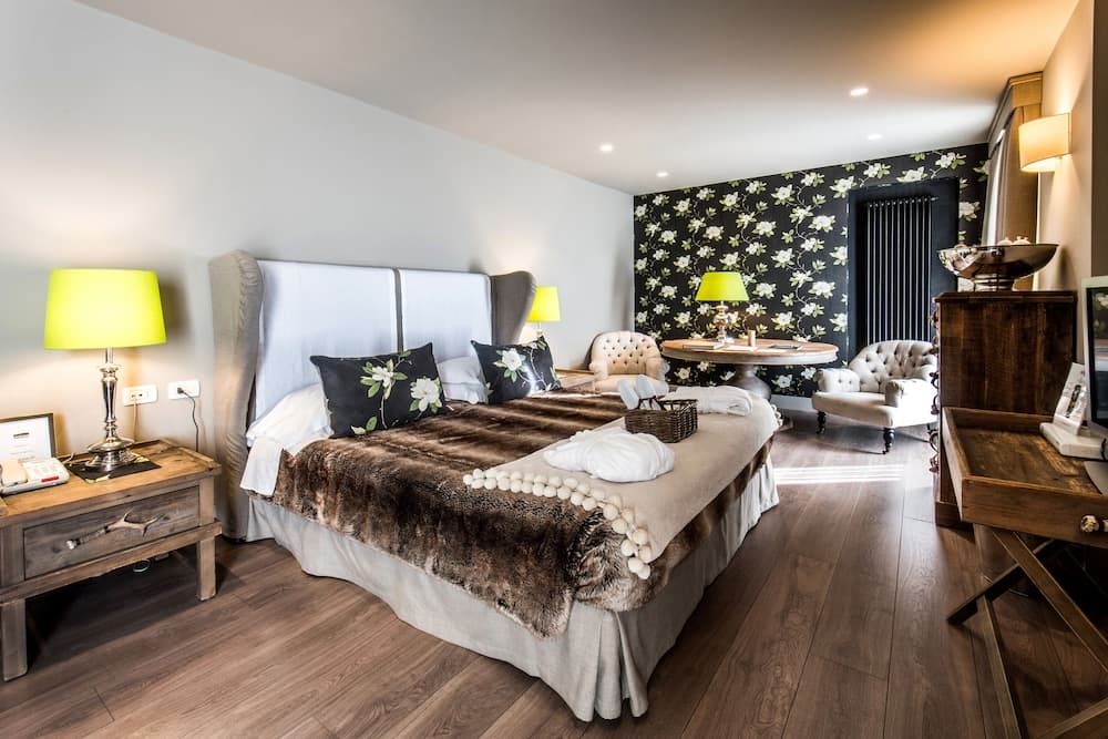 A cozy hotel room features a plush bed adorned with floral pillows and a fur throw, complemented by warm wooden floors, elegant armchairs, and vibrant green lamps, ideal for travelers seeking comfort and style.