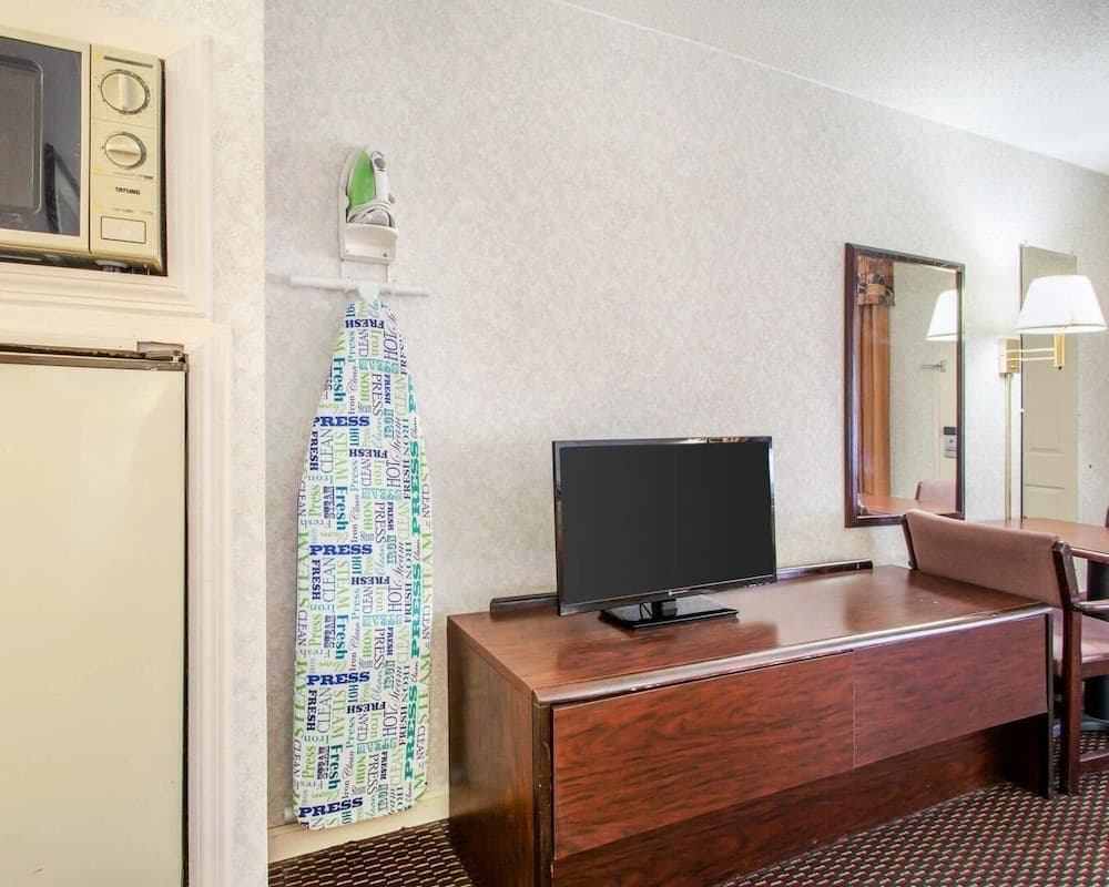 A cozy hotel room corner features a wooden desk with a flat-screen TV, a wall-mounted ironing board covered in colorful text, a microwave, and a mirror reflecting the serene ambiance, ideal for travelers seeking comfort and convenience.