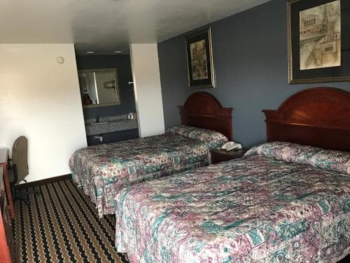 A cozy motel room features two floral-patterned beds against a blue wall, framed artwork, and a compact bathroom area, ideal for travelers seeking comfortable and budget-friendly accommodations.