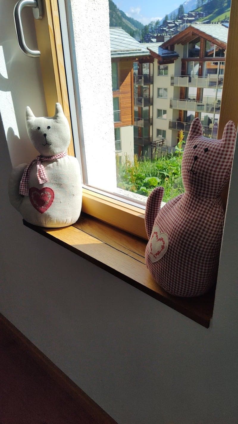 Two charming stuffed cats perch on a windowsill, gazing out at the picturesque alpine village of Zermatt, Switzerland, creating a cozy and inviting atmosphere perfect for travelers seeking a serene mountain retreat.
