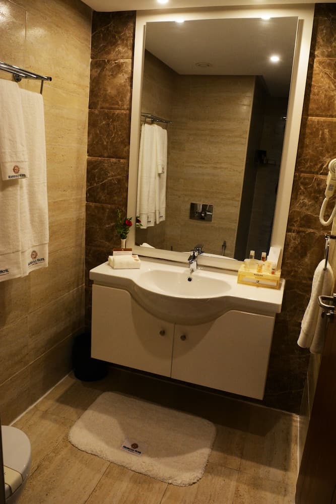 A cozy hotel bathroom features marble walls, a sleek white vanity with toiletries, a plush bath mat, and neatly hung towels, ideal for travelers seeking comfort and cleanliness.