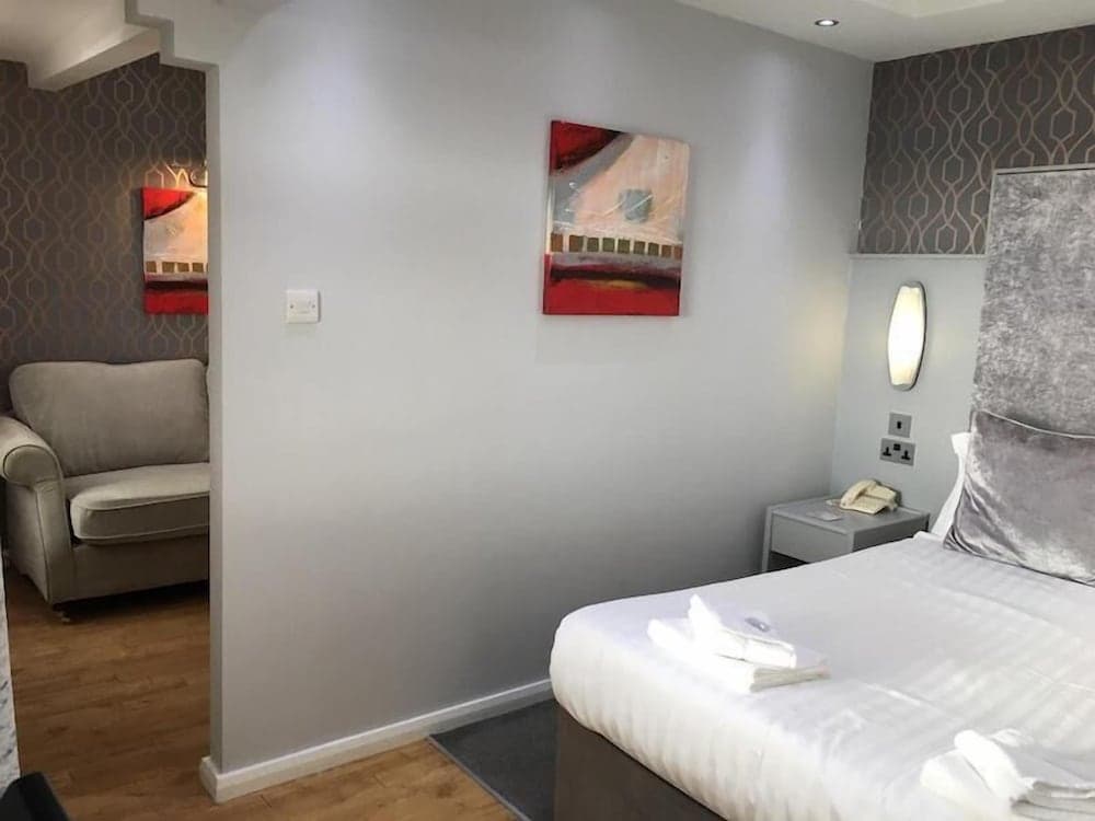 A cozy hotel room features a neatly made bed with white linens, modern artwork on gray walls, a comfortable armchair, and warm lighting, ideal for travelers seeking relaxation and comfort.