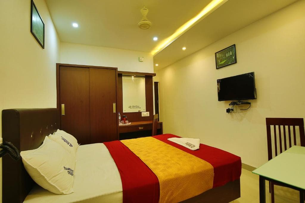 A cozy hotel room features a neatly made bed with red and gold linens, a small dining table, a mounted TV, and warm lighting, ideal for travelers seeking comfort and convenience.