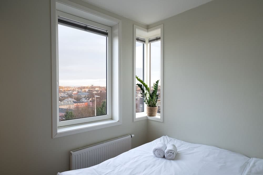 A serene bedroom with large windows offering a cityscape view, featuring a neatly made bed with rolled towels, a radiator, and a potted plant, ideal for travelers seeking a cozy urban retreat.