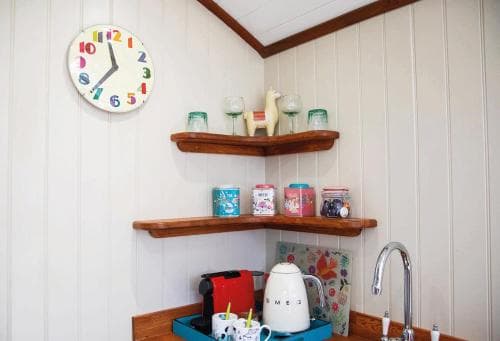 A cozy kitchen corner with wooden shelves displaying colorful mugs and glasses, a playful clock on the wall, and a charming kettle, perfect for a quaint getaway or a rustic cabin retreat.