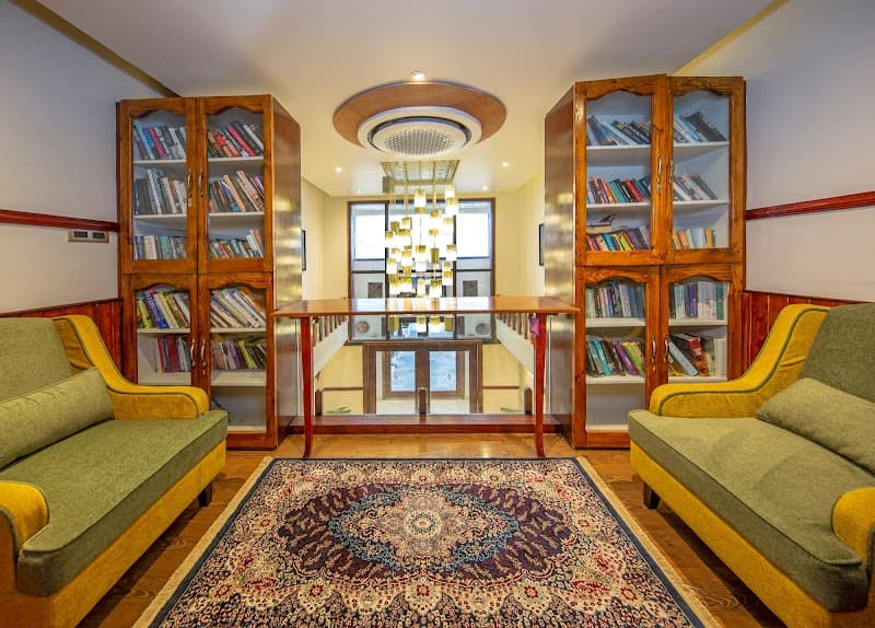 A cozy library lounge at an upscale resort features plush green sofas, wooden bookshelves filled with books, a vibrant patterned rug, and a modern chandelier, creating a serene atmosphere ideal for relaxation and leisure reading during a luxurious getaway.