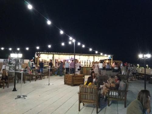 A lively rooftop bar scene at night, illuminated by string lights, features patrons enjoying drinks and conversation on wooden benches, ideal for social gatherings and evening outings.