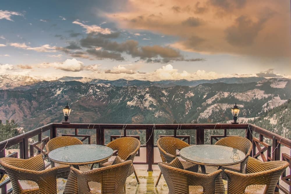 A serene mountain retreat features a cozy outdoor dining area with wicker chairs and tables overlooking snow-capped peaks under a dramatic sky, ideal for travelers seeking relaxation and breathtaking views.
