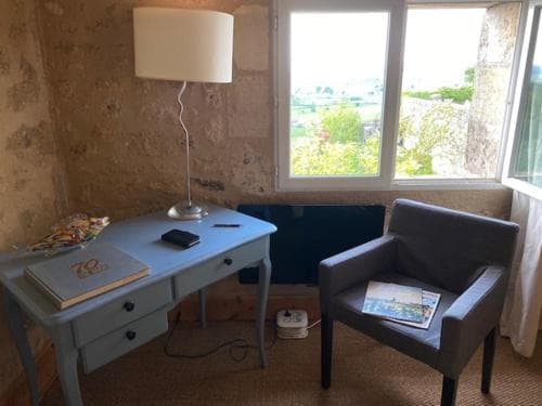 A cozy corner in a rustic room features a blue desk with books and a lamp, a gray armchair with magazines, and a window offering a serene view of greenery, ideal for travelers seeking a peaceful reading nook.