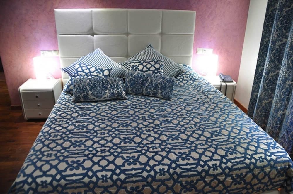 A cozy bedroom with a blue patterned bedspread, white tufted headboard, soft pink walls, and ambient bedside lamps creates an inviting atmosphere ideal for travelers seeking comfort and relaxation.
