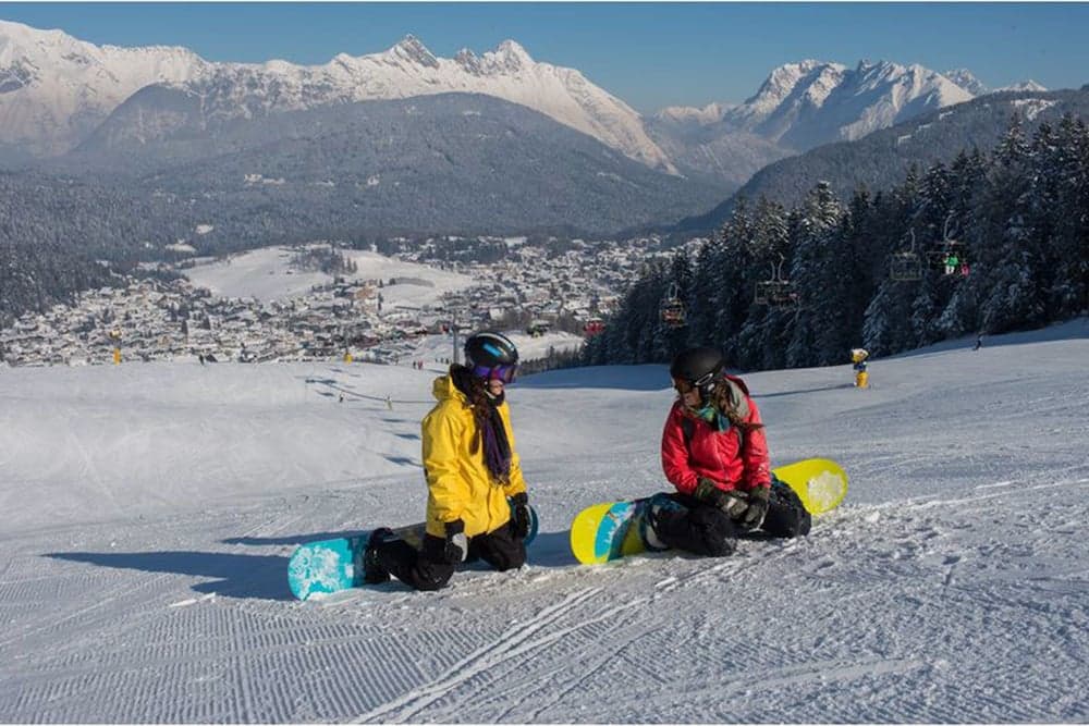 Two snowboarders rest on a sunny ski slope with breathtaking views of snow-capped mountains and a quaint village below, ideal for winter sports enthusiasts seeking relaxation and scenic beauty.