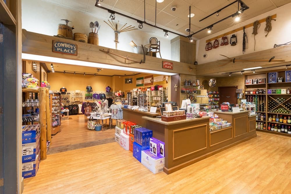 A cozy Western-themed store with wooden floors, rustic decor including barrels and lanterns, and shelves stocked with apparel and spirits, ideal for travelers seeking unique cowboy-inspired souvenirs and gifts.