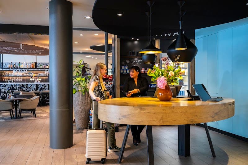 A traveler interacts with staff at the modern reception desk of an urban hotel in Amsterdam, featuring sleek decor, vibrant floral arrangements, and a cozy bar area, ideal for business or leisure travelers seeking a welcoming atmosphere.