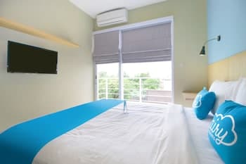 A cozy hotel room with a neatly made bed adorned with blue pillows, a bright window view of greenery, and modern amenities like a wall-mounted TV, ideal for travelers seeking comfort and relaxation.