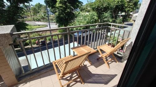 A cozy balcony with wooden chairs overlooks a serene street lined with lush greenery, perfect for travelers seeking relaxation in an urban retreat.