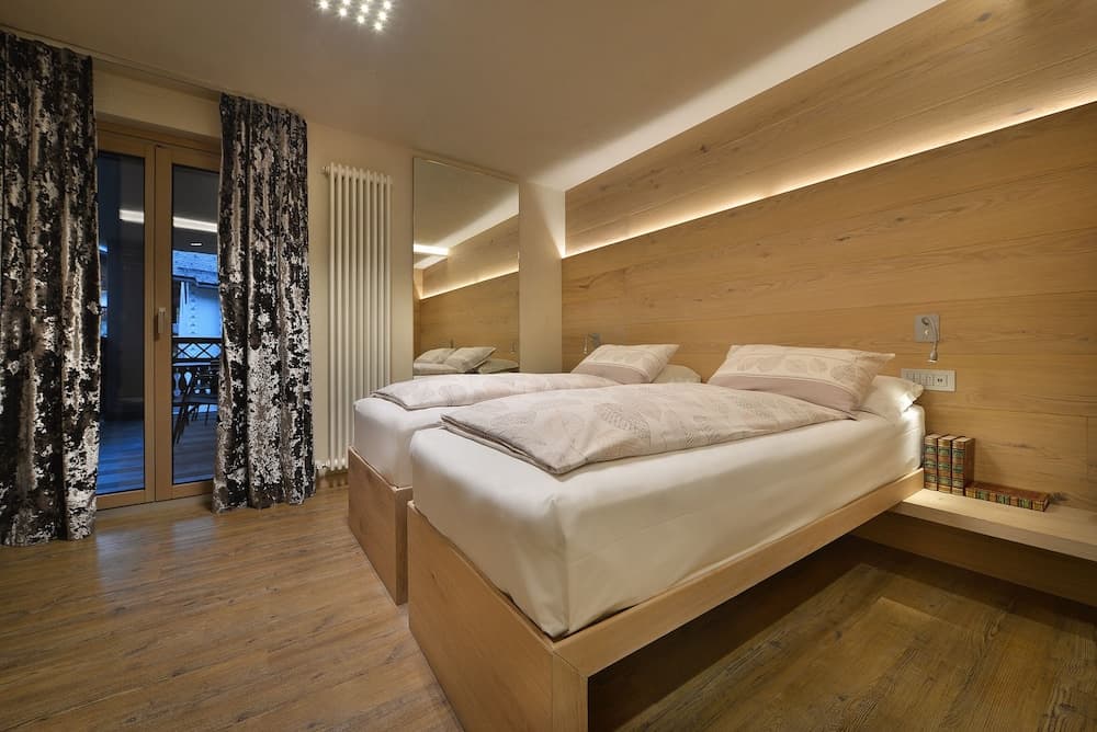 A cozy hotel bedroom with wooden walls and floors, featuring two neatly made beds, patterned curtains, and a balcony view, ideal for travelers seeking comfort and relaxation.