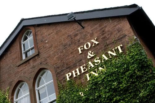 A charming brick building adorned with ivy displays the sign "FOX & PHEASANT INN," evoking a cozy, rustic pub atmosphere ideal for travelers seeking a quaint countryside retreat.
