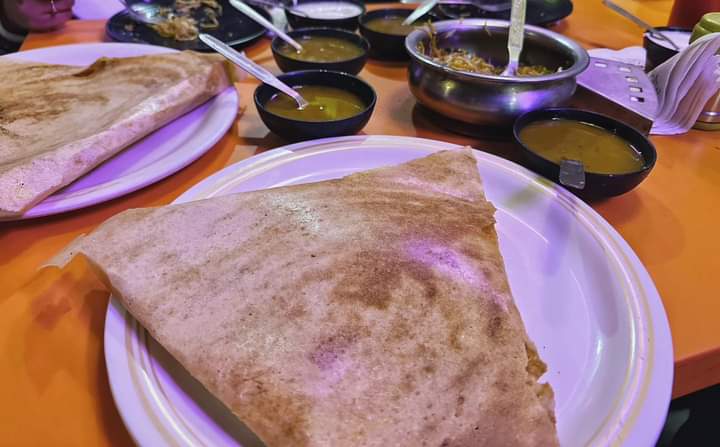 A cozy Indian restaurant scene featuring dosas on white plates surrounded by small bowls of chutneys, set on an orange table, ideal for travelers seeking authentic South Indian cuisine in a bustling bazaar area.