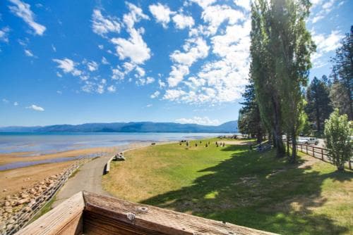 A serene lakeside scene with a grassy path, tall trees, and distant mountains under a bright blue sky, ideal for tranquil outdoor activities like hiking or picnicking.