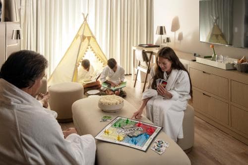 A cozy hotel room scene captures a family enjoying board games and reading in bathrobes, featuring a playful teepee tent and warm lighting, ideal for family-friendly travel experiences.