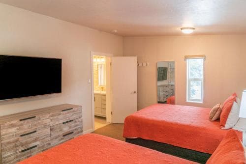 A cozy hotel room features two beds with vibrant orange bedding, a wooden dresser with a mounted TV, and a bathroom peeking through an open door, ideal for travelers seeking comfort and convenience.