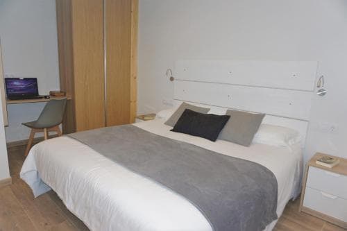 A cozy hotel room features a neatly made bed with gray accents, a small desk with a laptop, wooden furnishings, and a minimalist design, ideal for travelers seeking comfort and simplicity.