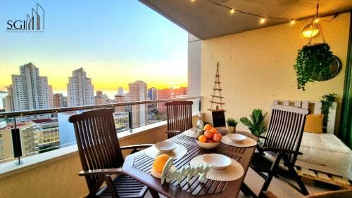 A cozy balcony adorned with string lights, plants, and a dining setup overlooks a stunning cityscape at sunset, ideal for urban retreats and romantic getaways.