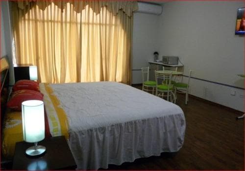 A cozy hotel room features a neatly made bed with white linens, a small dining area with green chairs, warm yellow curtains, and ambient lighting, ideal for travelers seeking comfort and functionality.