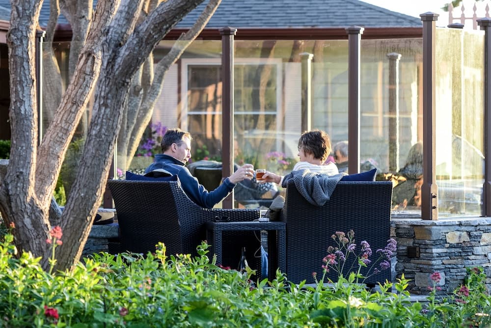 A serene backyard patio scene features two people enjoying drinks amidst lush greenery and blooming flowers, framed by modern wicker furniture and glass-enclosed structures, ideal for travelers seeking peaceful outdoor relaxation spots.