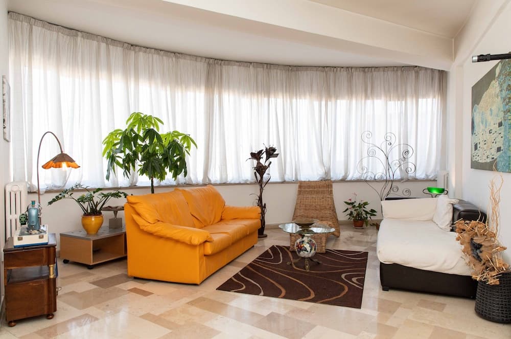A cozy living space with a vibrant orange sofa, tiled floor, and large curtained windows creates a warm and inviting atmosphere, ideal for travelers seeking a comfortable apartment stay.