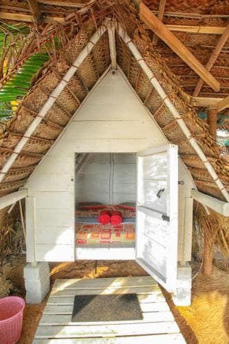 A cozy thatched-roof hut with an open door revealing vibrant red cushions inside invites travelers to unwind in a serene tropical retreat.