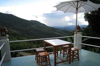 A serene balcony overlooking lush green mountains features a wooden table with stools under a white umbrella, ideal for travelers seeking a peaceful retreat spot.
