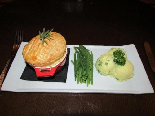 A cozy dining scene features a golden puff pastry pot pie served with vibrant green beans and creamy mashed potatoes garnished with parsley on a white plate, ideal for travelers seeking a comforting meal in a restaurant setting.