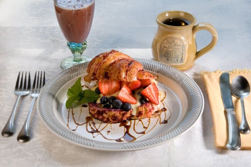 A cozy breakfast scene featuring a plated croissant dessert topped with fresh berries and syrup, accompanied by a mug of coffee and a chocolate milkshake, set on an elegant tablecloth, perfect for a relaxing morning getaway.