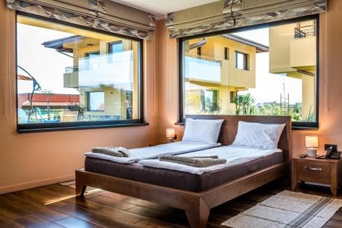 A cozy hotel room with a neatly made bed, large windows offering a view of modern buildings and greenery, exudes a serene and inviting atmosphere ideal for travelers seeking relaxation.