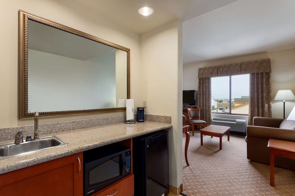 A cozy hotel suite features a compact kitchenette with a sink, microwave, and fridge under a large mirror, leading to a warmly lit living area with comfortable seating and a view through the window, ideal for travelers seeking convenience and comfort.