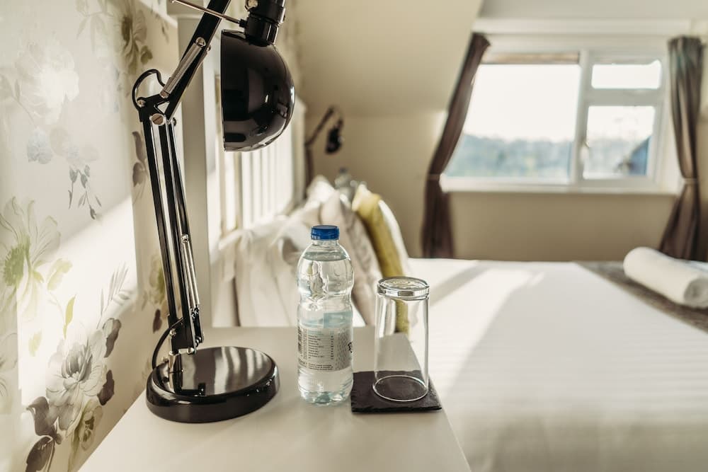 A cozy hotel room with floral wallpaper exudes tranquility, featuring a bedside table with a sleek black lamp, a water bottle, and a glass, perfect for a relaxing stay.