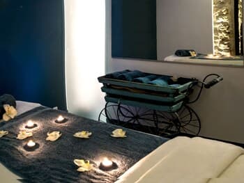 A serene spa room features a massage table adorned with floating candles and white flowers, complemented by a vintage carriage holding towels, creating a tranquil atmosphere ideal for relaxation and wellness retreats.