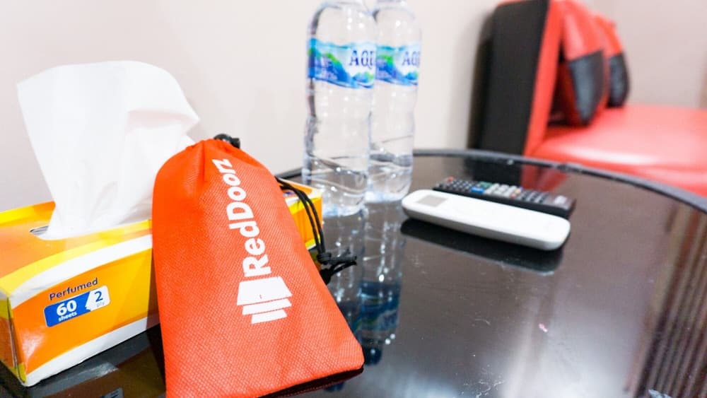 A cozy hotel room table setup featuring an orange RedDoorz pouch, tissue box, water bottles, remote controls, and a vibrant red chair, ideal for travelers seeking comfort and convenience.