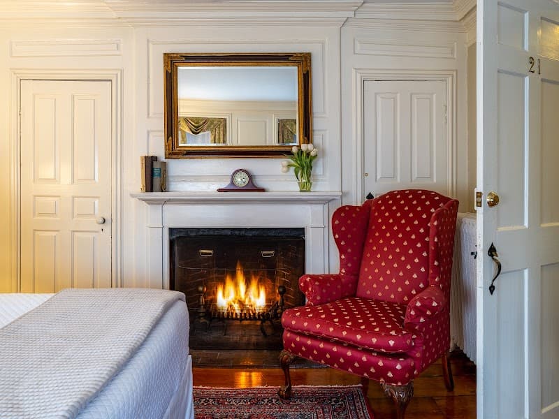 A cozy guest room at the Harbor Light Inn features a warm fireplace, elegant red armchair, and classic decor, perfect for travelers seeking a charming and inviting stay in Marblehead, MA.