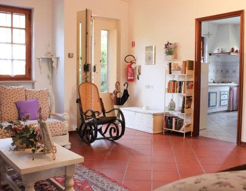 A cozy living room with terracotta flooring, a rocking chair, bookshelves, and an open doorway to a kitchen, evokes a warm, inviting atmosphere perfect for a relaxing vacation rental experience.