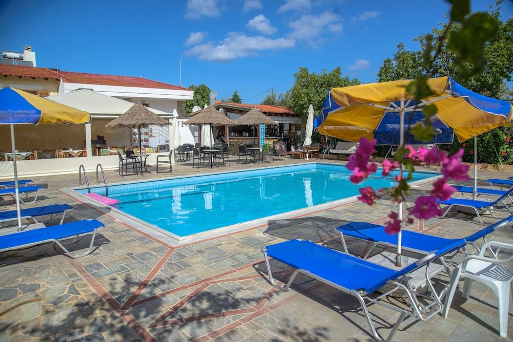 A serene resort pool area features vibrant blue lounge chairs under colorful umbrellas, surrounded by lush greenery and a clear blue sky, ideal for relaxing vacations.
