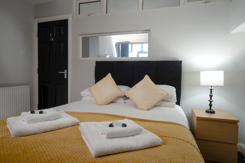 A cozy bedroom with a neatly made bed adorned with yellow bedding and white towels, featuring a black headboard, wooden bedside table with a lamp, and a small window above the bed, ideal for travelers seeking a comfortable and inviting accommodation.