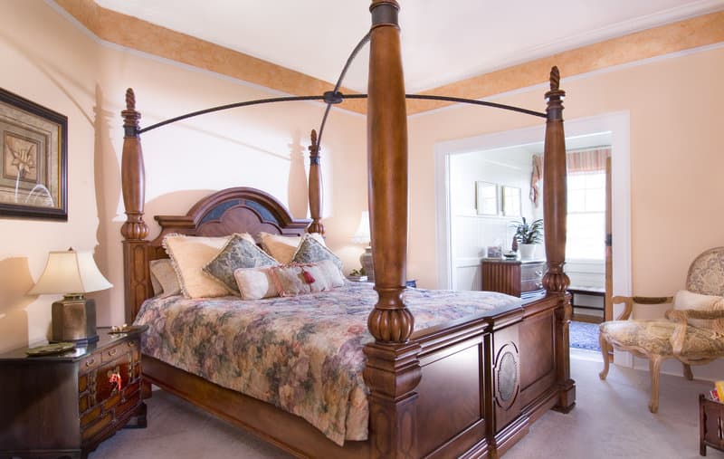 A cozy hotel room features a grand four-poster bed adorned with floral bedding, warm-toned walls, elegant furnishings, and soft lighting, ideal for travelers seeking a luxurious and relaxing retreat.