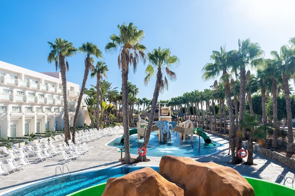 A vibrant resort pool area surrounded by lush palm trees under a clear blue sky, featuring playful water slides and lounge chairs, ideal for family vacations seeking relaxation and fun.