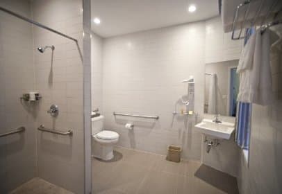 A clean, accessible hotel bathroom features tiled walls, safety bars, a shower, toilet, sink, and neatly hung towels, ideal for travelers seeking comfort and convenience.