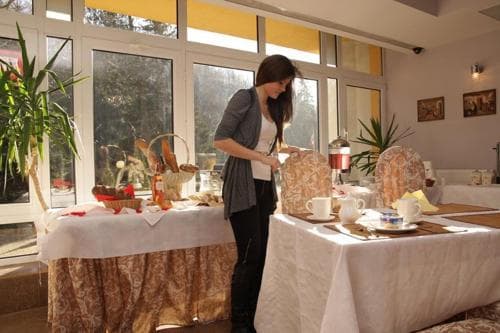 A serene indoor dining area bathed in natural light from large windows features a woman arranging a cozy breakfast spread on elegant tables, ideal for a relaxing retreat or wellness getaway.